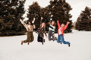 Four friends jump joyfully in the snow with evergreen trees in the background.