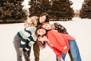 A group poses playfully together outdoors in winter clothing against a backdrop of evergreen trees.