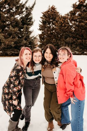 Four friends pose together in winter clothing against a backdrop of snow-covered evergreen trees.