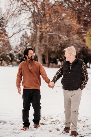 A couple holds hands and walks together through a snowy winter landscape with brown autumn trees in the background.