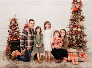Family poses in front of two decorated Christmas trees with red and gold ornaments and wrapped presents on the floor.