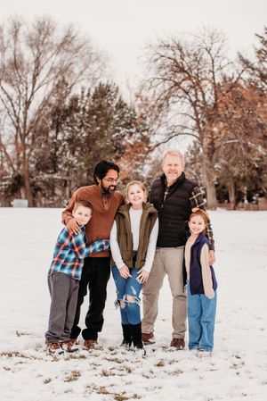 A group stands together smiling in a snowy park setting during winter with bare trees in the background.