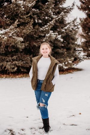 A person wearing a brown vest and jeans plays in the snow with evergreen trees in the background.