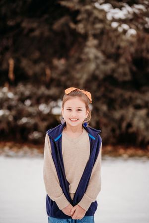 A person wearing a navy vest and beige sweater smiles during a winter photo session in the snow.