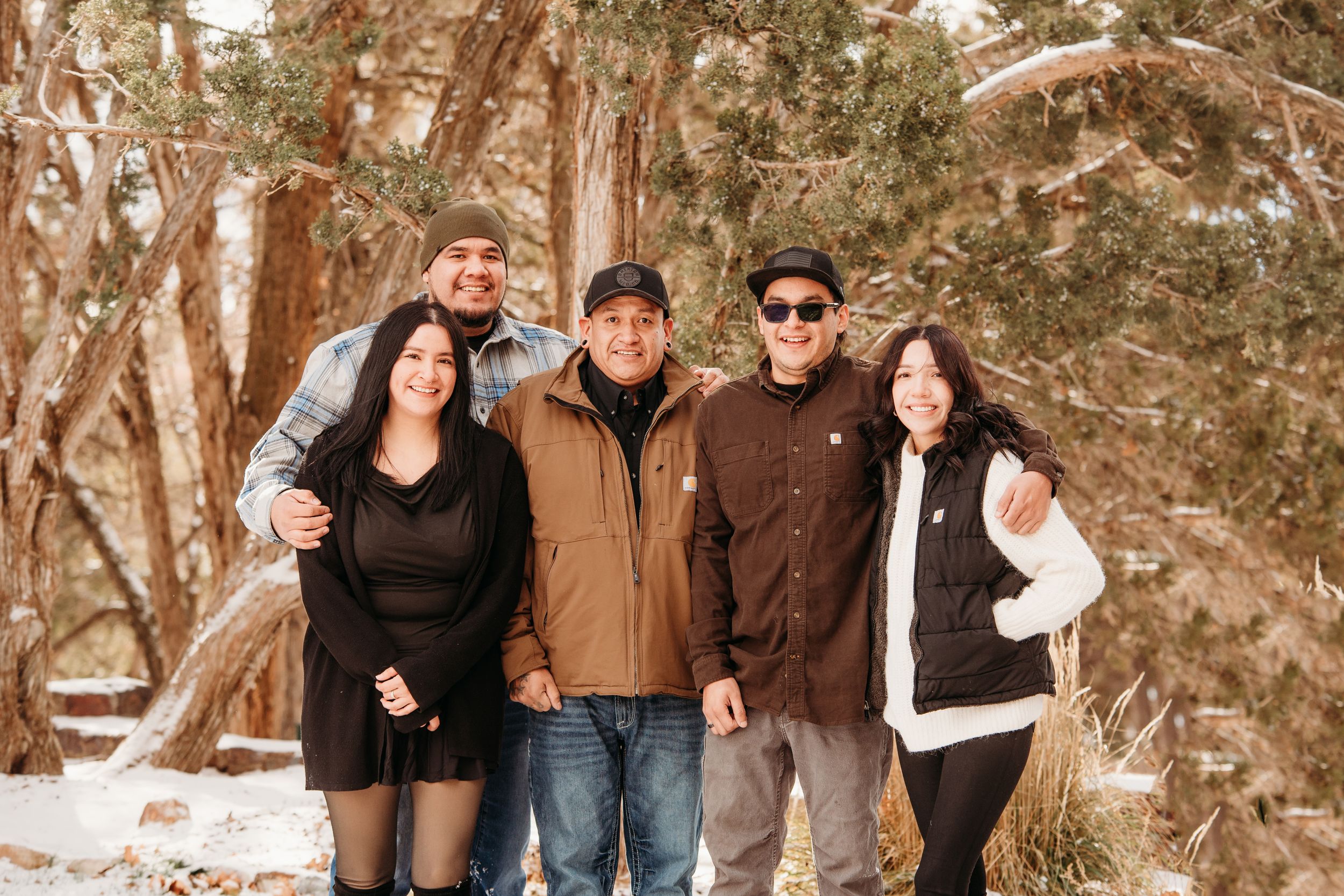 A group of people pose together outdoors in winter clothing against a snowy forest backdrop.