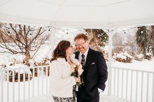A romantic winter wedding moment on a snow-covered gazebo with white railing and bare trees in the background.