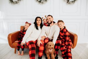 Family wearing matching red and black buffalo plaid pajamas pose together on brown leather couch for holiday photos.