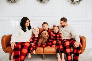 A family wearing matching red and black buffalo plaid pajamas sits together on a brown leather couch.