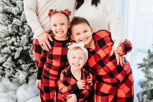 Family wearing matching red and black buffalo plaid pajamas poses in front of snowy Christmas trees for holiday photos.