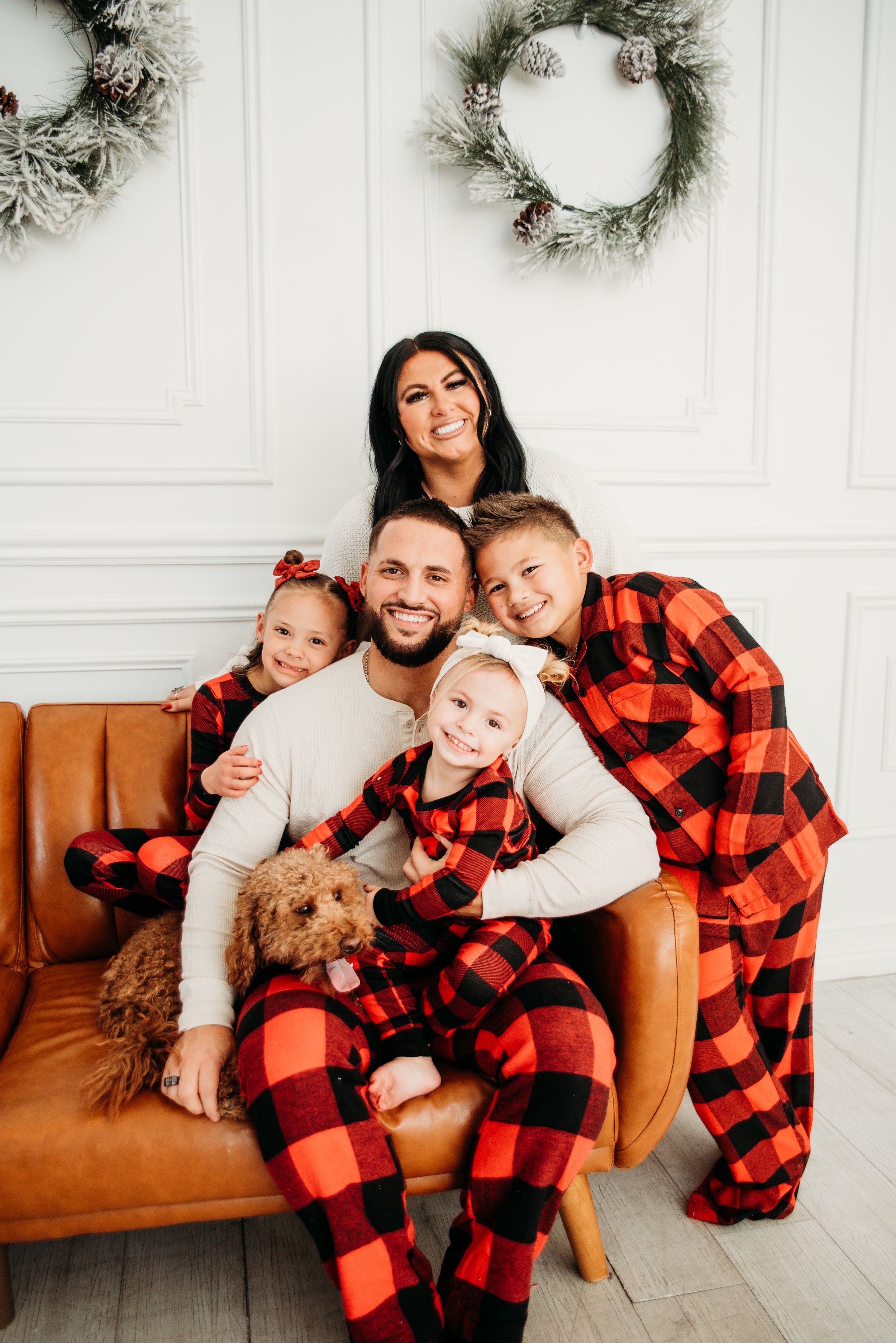 A family wearing matching red buffalo plaid pajamas poses together on a brown leather couch with holiday decor in background.