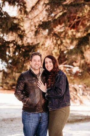A couple poses together in a snowy winter setting with evergreen trees in the background during an engagement photo session.