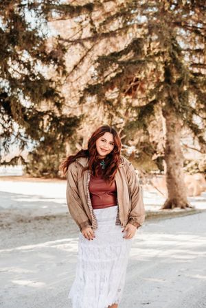 A person in a brown jacket and burgundy top poses playfully in a snowy winter setting with evergreen trees.