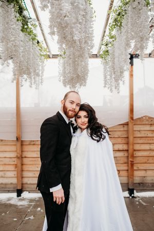 A couple stands under cascading white wisteria flowers at their winter wedding ceremony.