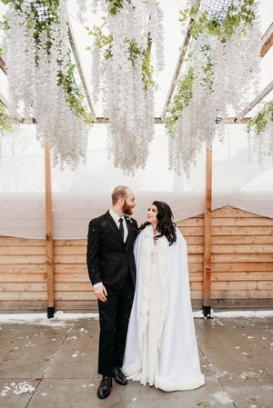 A romantic wedding couple stands beneath cascading white wisteria flowers hanging from a wooden pergola.