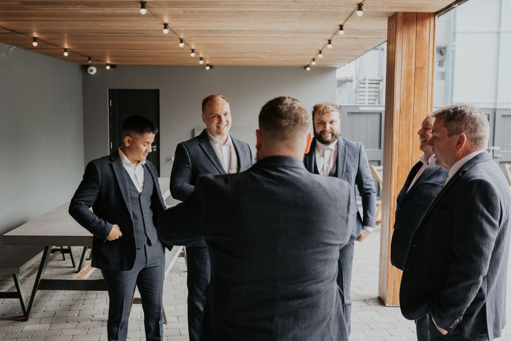 Group of groomsmen in grey suits chatting on an outdoor terrace with string lights.