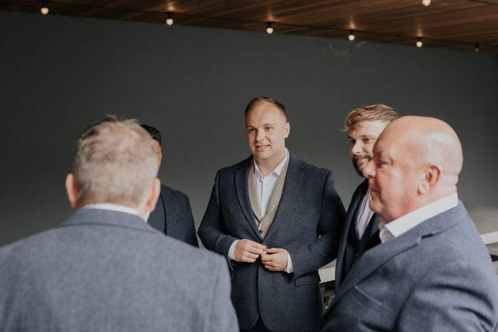 Groomsmen in grey suits smiling and conversing inside a modern venue with grey walls.