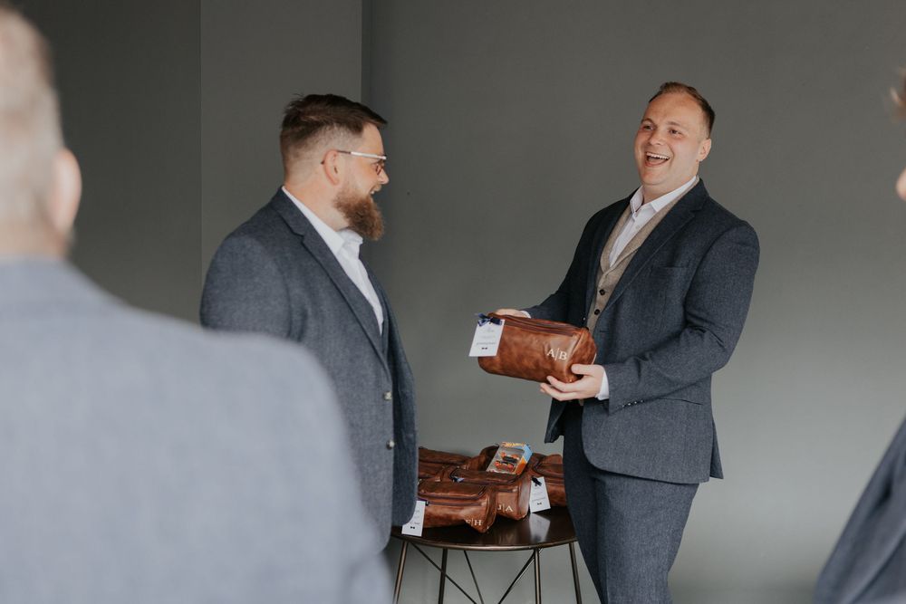 Two groomsmen in grey suits smiling while one holds a brown leather toiletry bag gift.