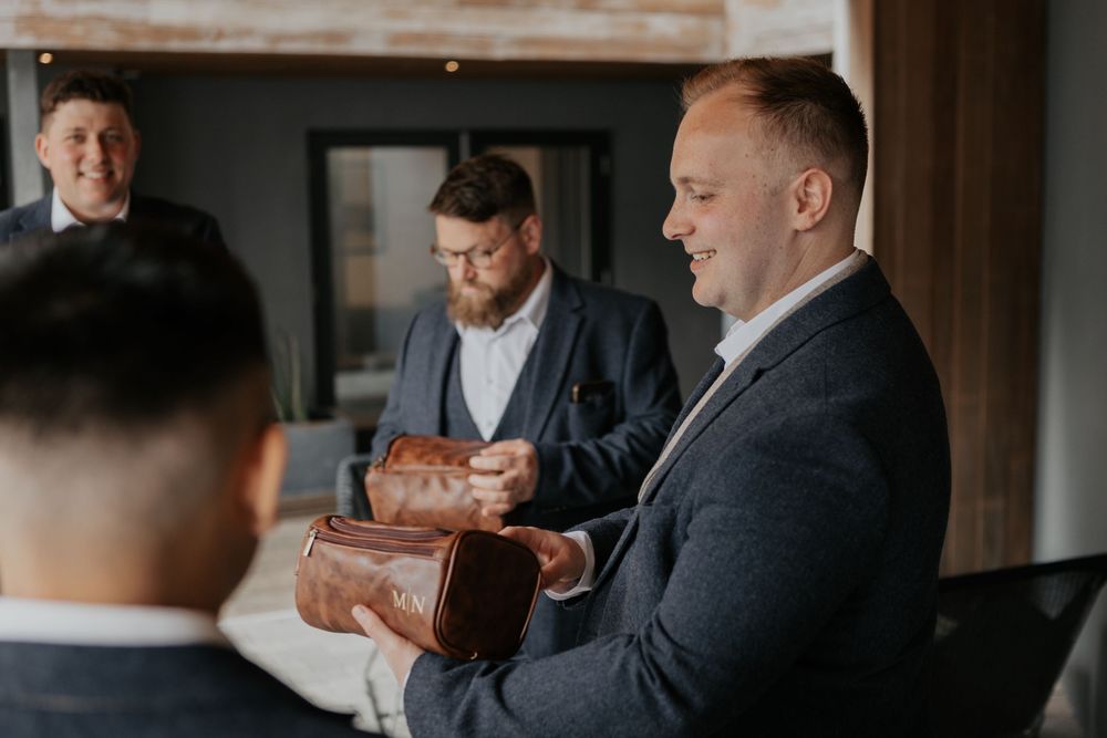 Groom presenting a brown leather toiletry bag gift to a smiling groomsman indoors.