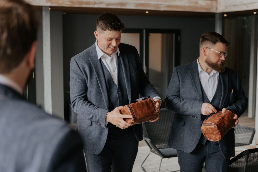 Two groomsmen in grey suits opening brown leather toiletry bag gifts and smiling.