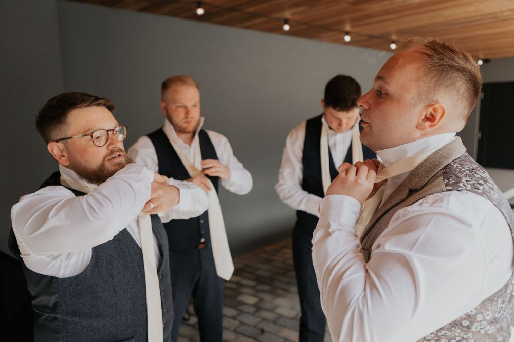 Groomsmen in white shirts and suspenders helping each other adjust ties before the wedding.