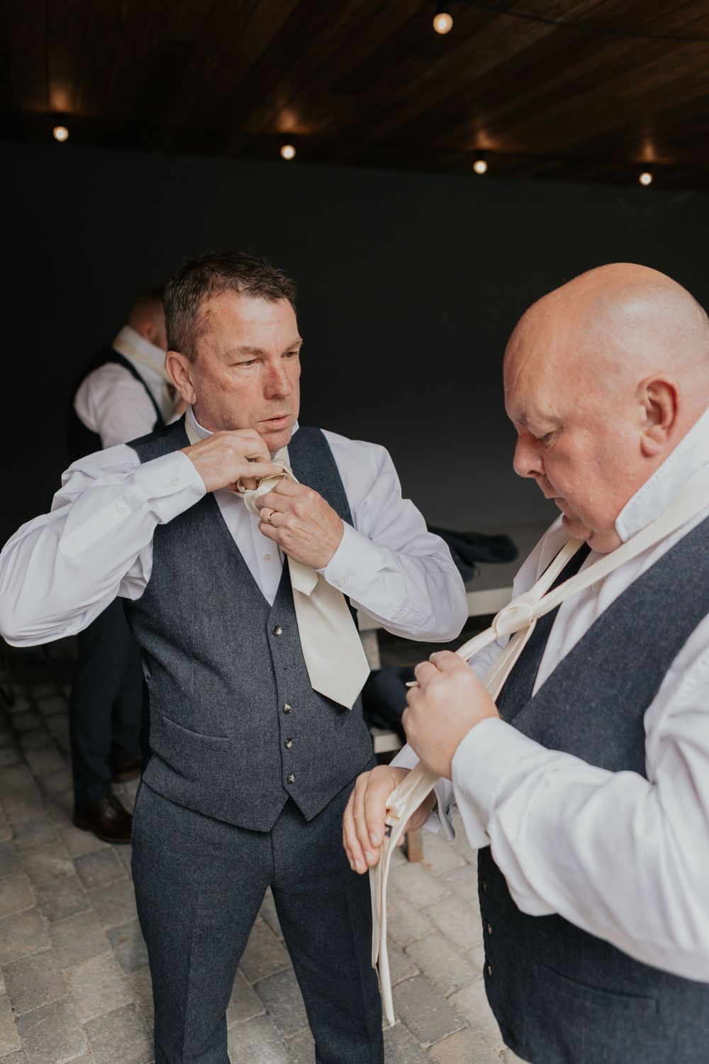 Groomsmen helping each other adjust cream ties and waistcoats while getting ready for a wedding.