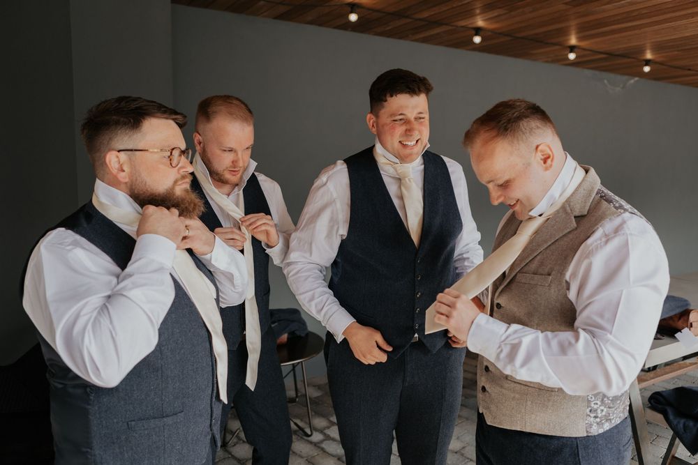Four groomsmen smiling and adjusting ties and waistcoats while getting dressed for a wedding.