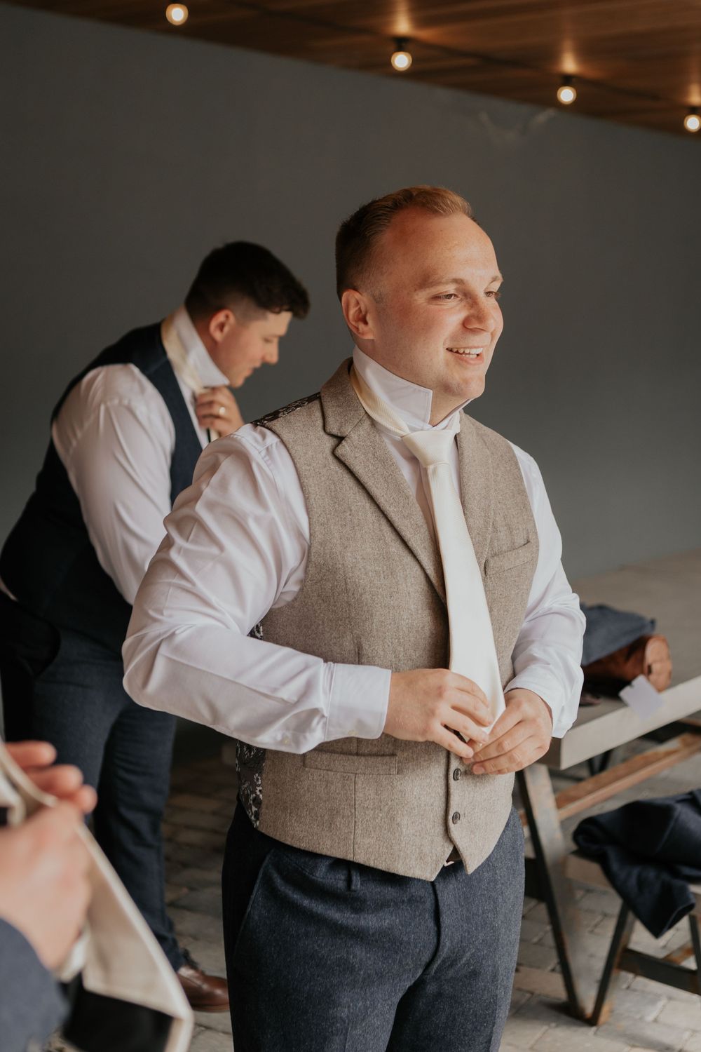 Smiling groom in beige tweed waistcoat buttoning up while groomsmen get ready in the background.