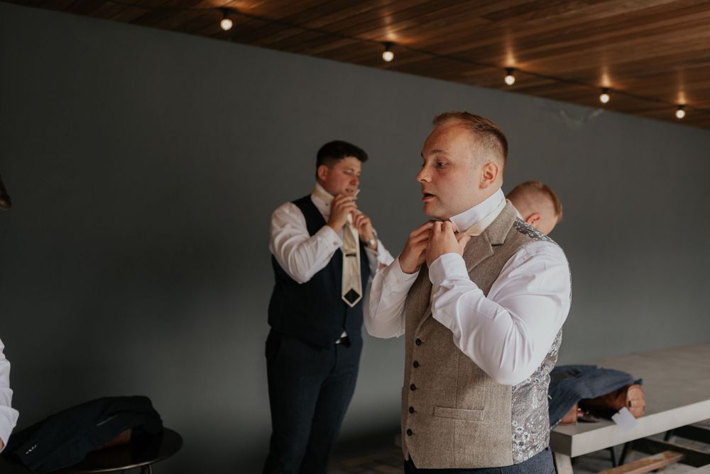 Groomsmen in tweed waistcoats adjusting ties and preparing for a wedding ceremony indoors.