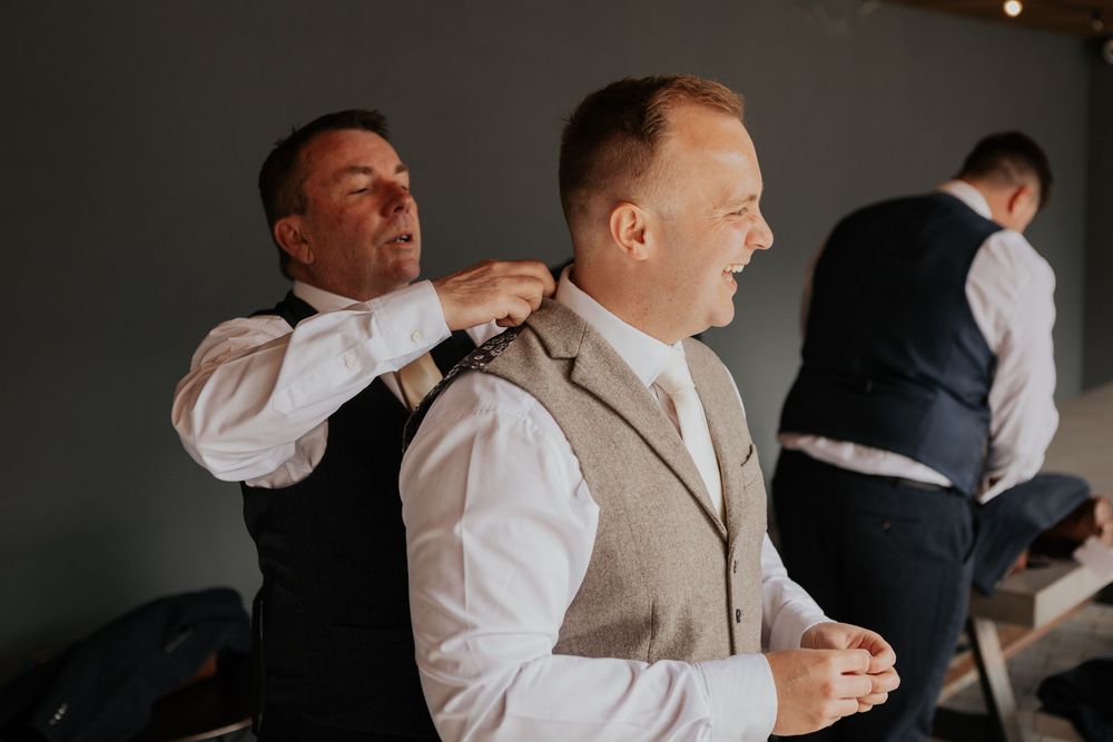 Groomsman helping adjust collar of man in beige tweed waistcoat during wedding preparations.