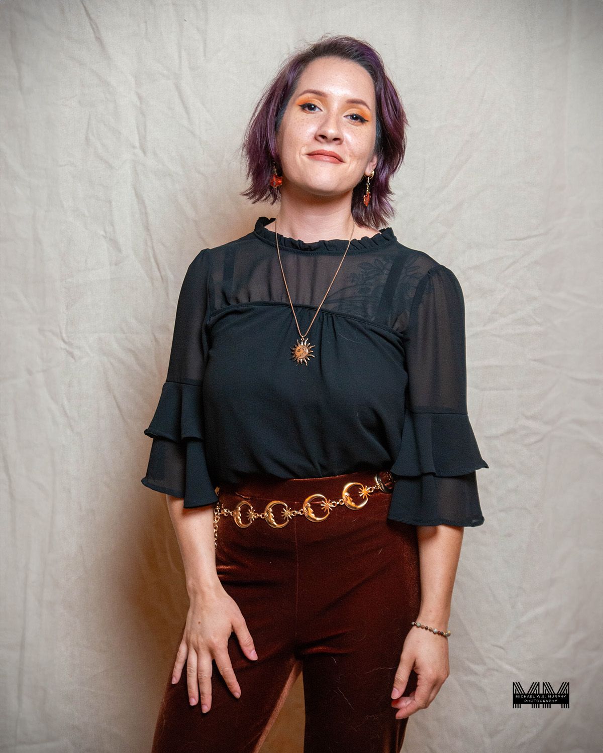 Portrait of person in black ruffled blouse with ornate belt against light background.