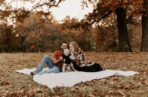 Wide landscape photo showing a family gathering on a white blanket among autumn leaves.