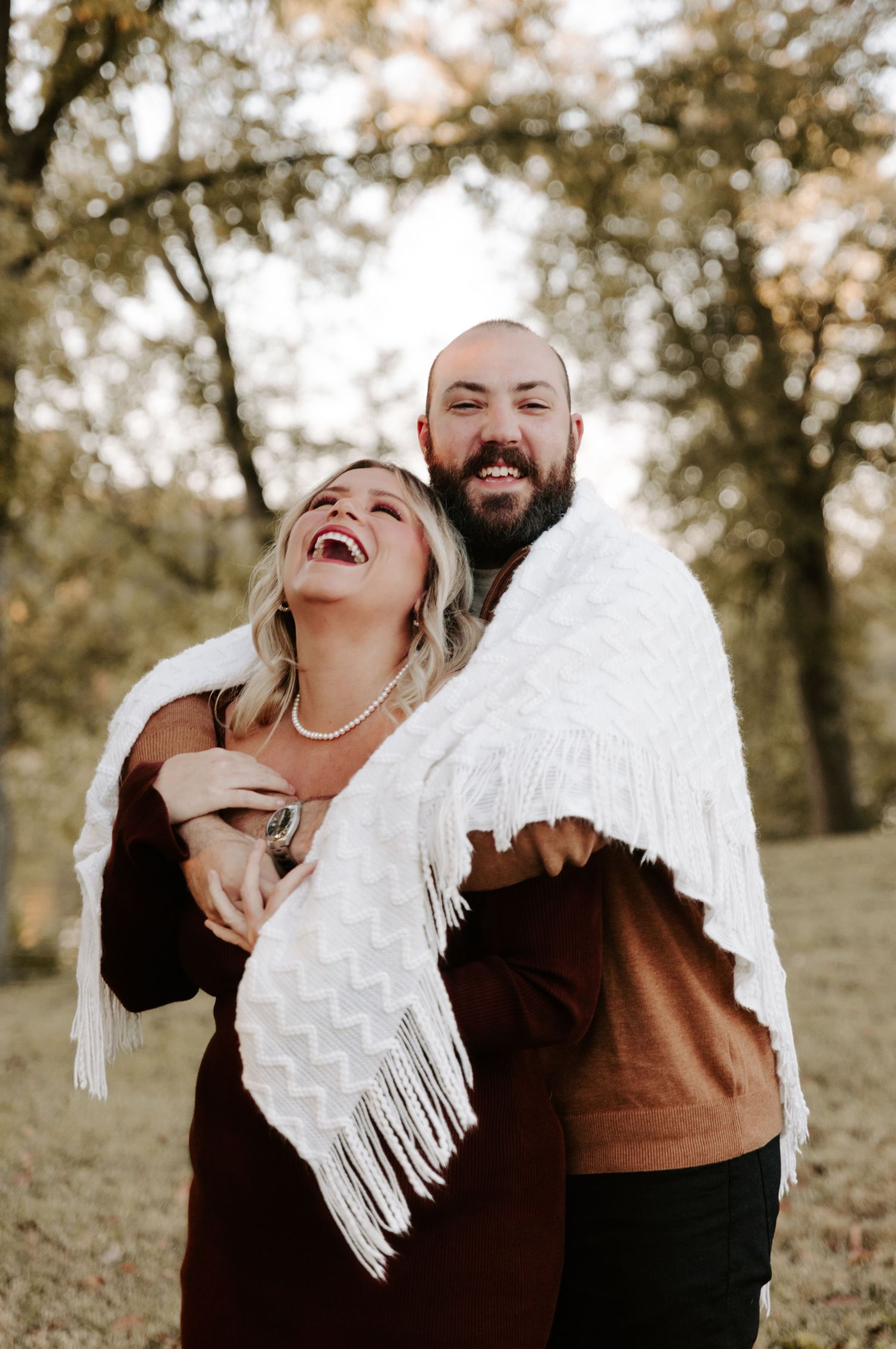 Joyful couple sharing laughter while wrapped in a white fringed blanket outdoors.