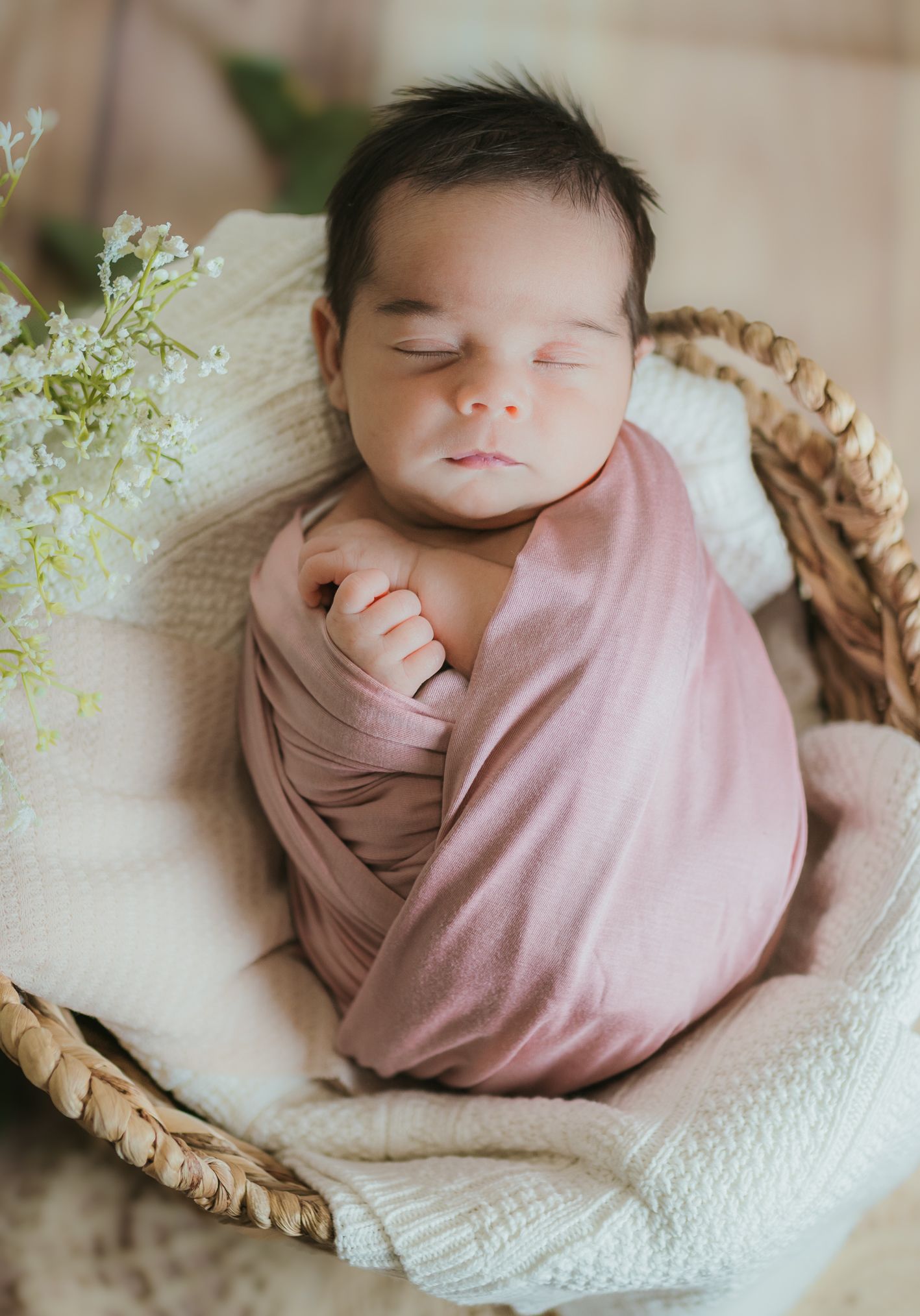 Sleeping newborn wrapped in soft pink swaddle lying in woven basket with delicate white flowers.Colorado Springs Photographer