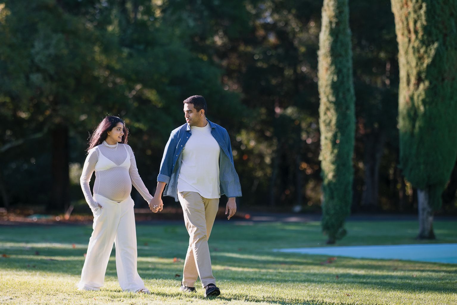 Pregnant couple holding hands and walking through sunny green park in coordinating white and blue casual outfits.