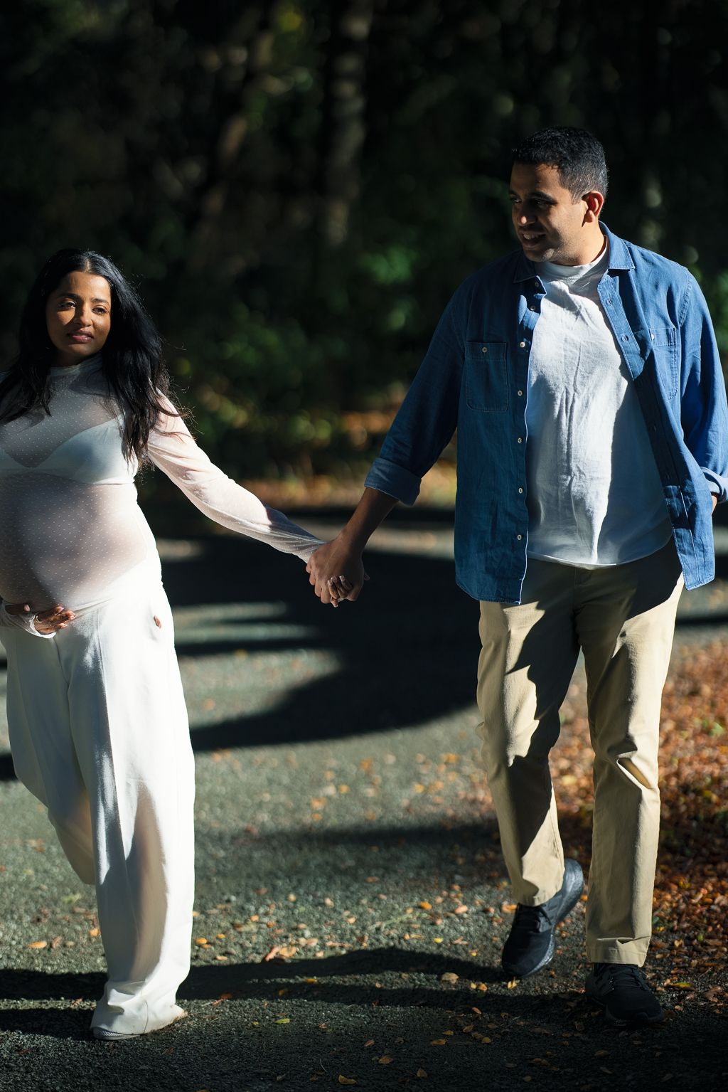 Maternity couple holding hands on sunlit path, woman in white sheer gown and man in blue shirt and khakis.