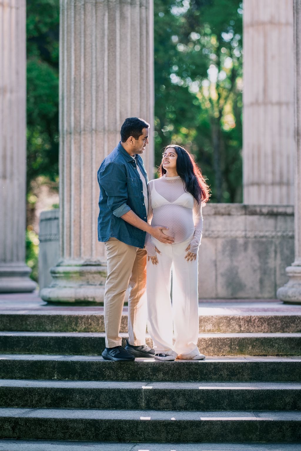 Expecting couple sharing tender moment on stone steps near classical columns, man gently touching pregnant belly.