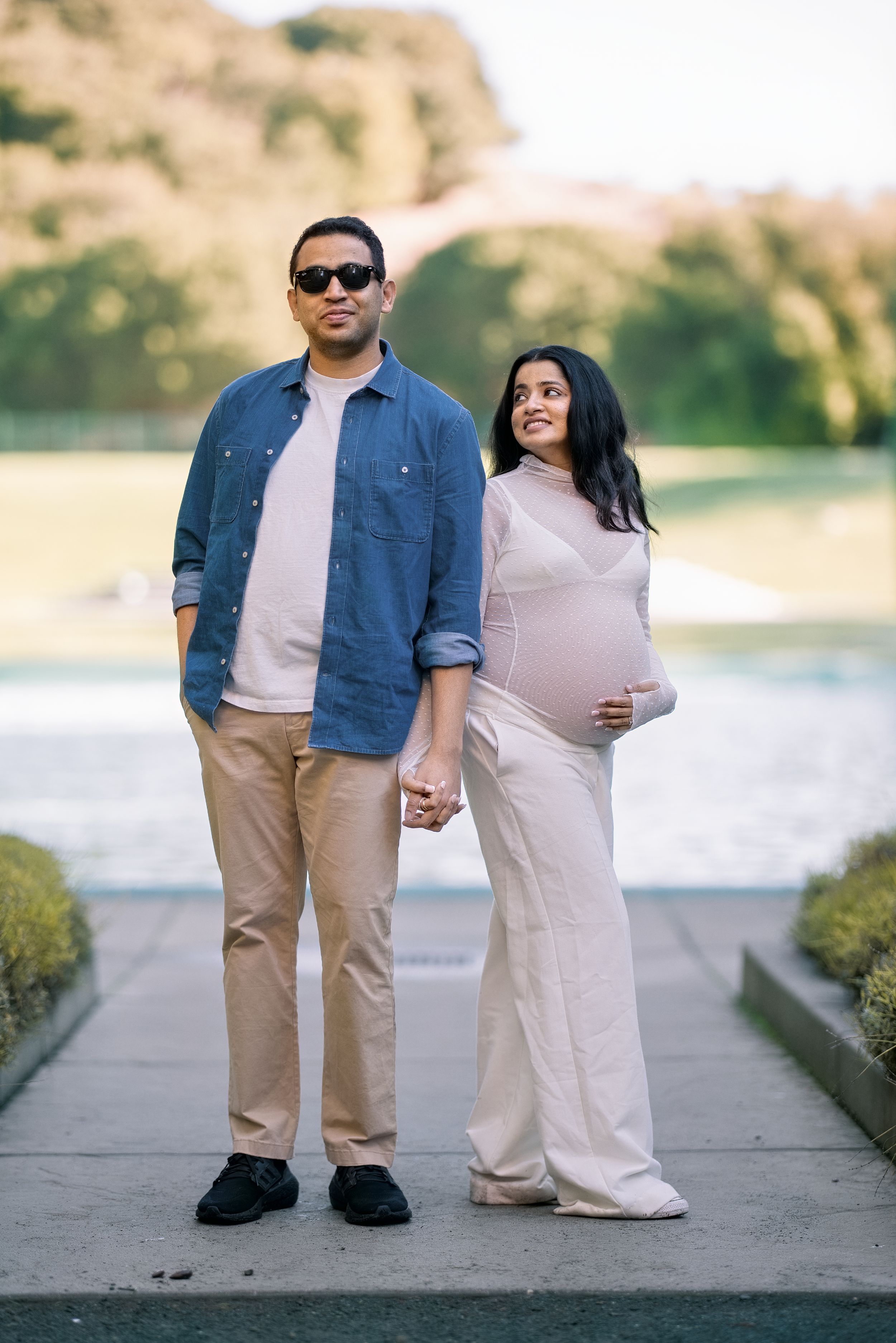 Maternity portrait of stylish couple holding hands outdoors, woman in white jumpsuit cradling baby bump at sunset.