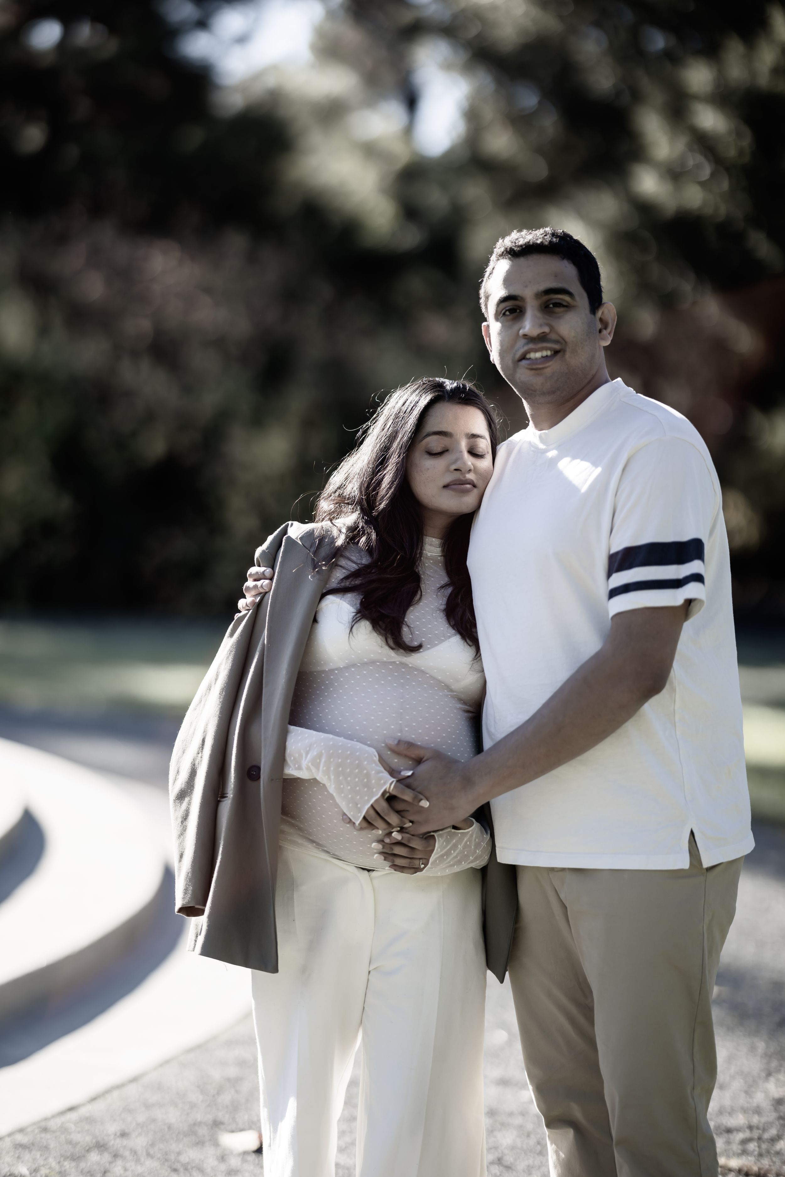 Maternity photo of couple in sunny park, man embracing pregnant woman from behind in soft natural light.