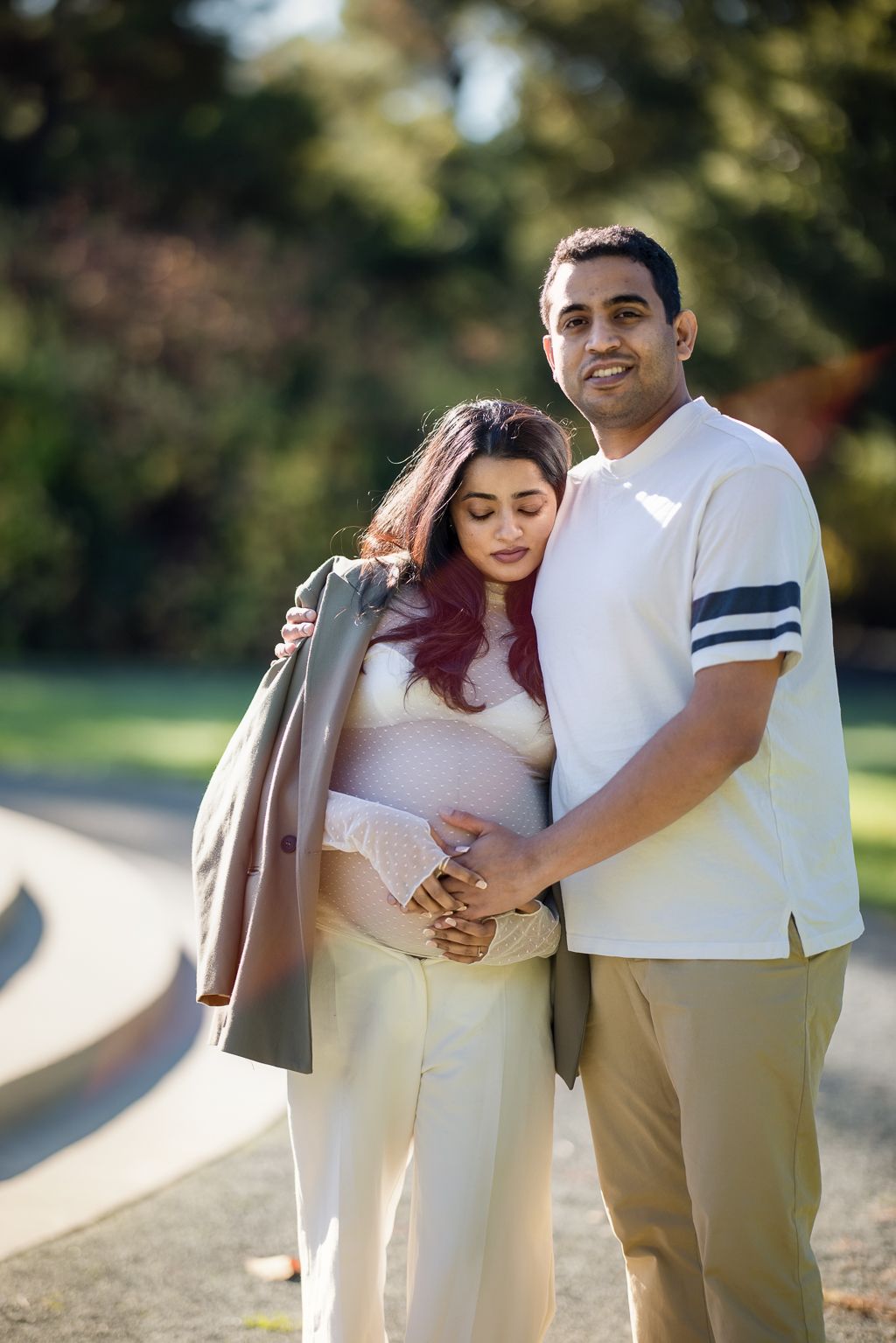 Expecting couple embracing outdoors in park, man in white tee holding pregnant woman in white outfit and gray jacket.