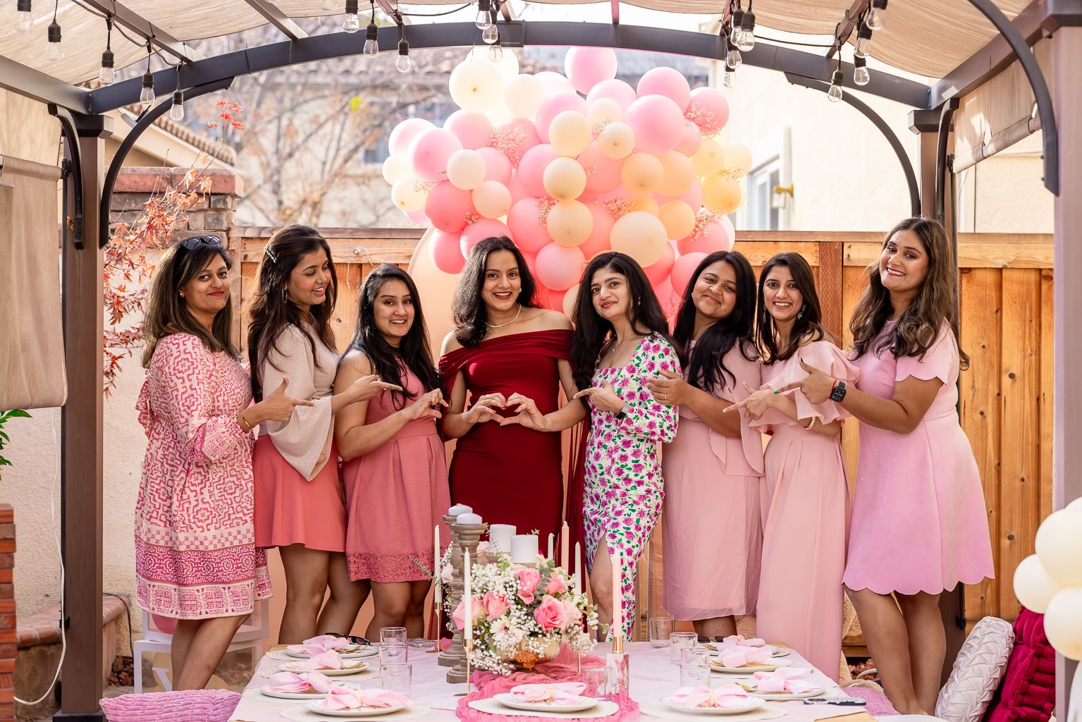 Pregnant woman in red dress surrounded by friends in pink at outdoor baby shower with balloons.