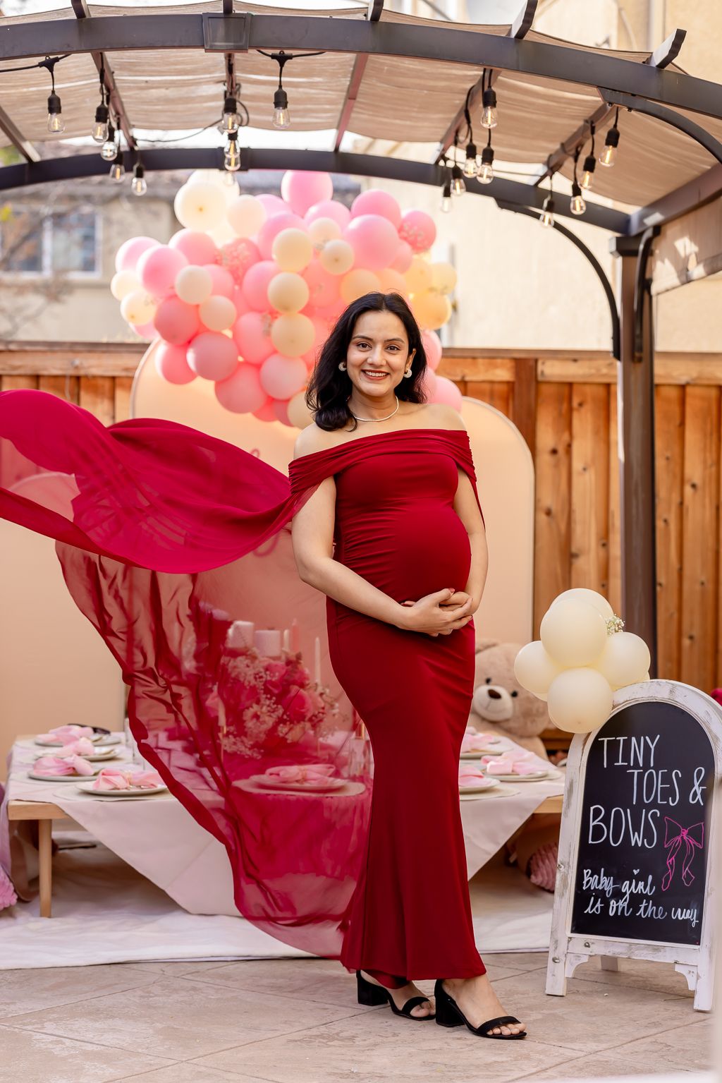 Pregnant woman in red off-shoulder gown poses at Tiny Toes and Bows baby shower with balloon arch.