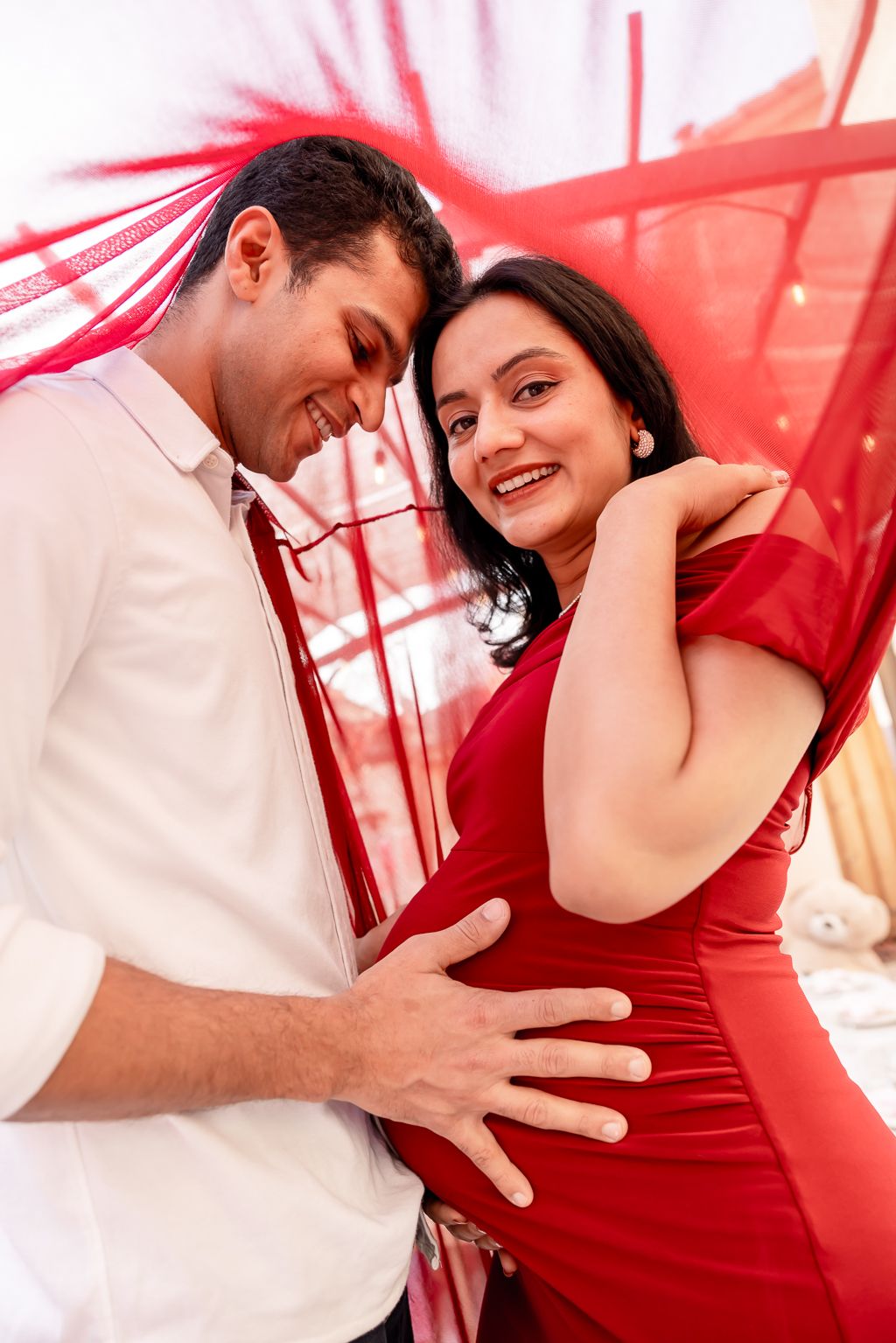 Smiling couple in maternity photoshoot, man touching baby bump, woman in red off-shoulder dress, red fabric backdrop.