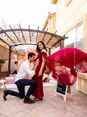 Pregnant woman in red gown with flowing fabric at baby shower as partner kneels kissing her belly.