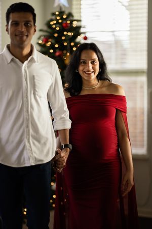 Smiling pregnant couple holding hands in front of Christmas tree, woman wearing red maternity dress.