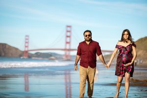 Pregnant couple walking hand-in-hand on Baker Beach, Golden Gate Bridge backdrop, woman in floral maternity dress.