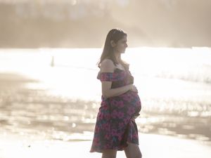 A pregnant woman in a floral dress walks along a sunlit beach with glowing backlight surrounding her silhouette.