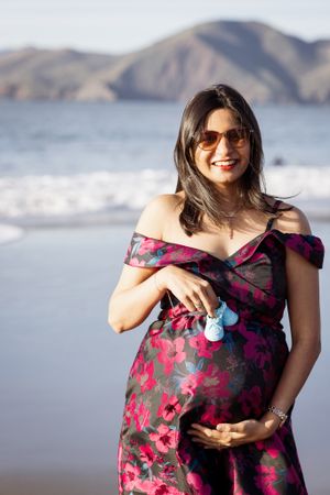 Smiling pregnant woman in floral dress holding baby shoes on beach, sunglasses, mountains in background.