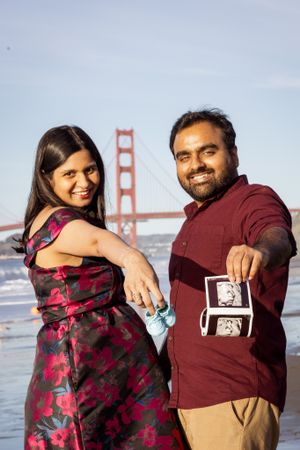 A smiling couple stands at Baker Beach holding baby shoes and ultrasound photos with the Golden Gate Bridge behind them.