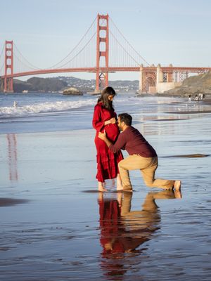 A man kneels before his pregnant partner in a red dress on a reflective beach with the Golden Gate Bridge backdrop.