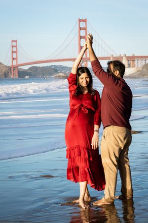 Couple dancing on Baker Beach with Golden Gate Bridge behind them, woman in red maternity dress, sunny day.